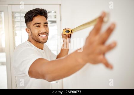 My picture frame will fit here perfectly. a handsome young man standing alone in his home and using measuring tape to measure a wall. Stock Photo