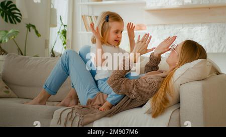 Happy parent Caucasian mom mother with cute kid daughter child girl baby playing patty cake game at home clapping hands lying on sofa couch at home Stock Photo