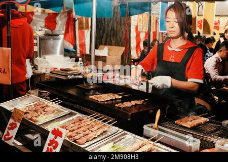 Tokyo, Japan - November 1, 2022 : Local Japanese street food at night market in Tokyo, Japan.  woman grilling yakitori pork and chicken at street food Stock Photo