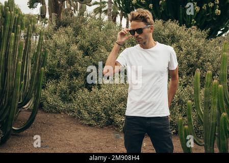 Blond attractive hipster guy  wearing white empty t-stirt and  sunglasses is standing in the garden near a big cactus. Stock Photo