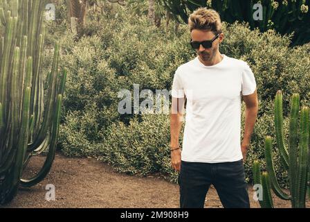 Blond attractive hipster guy  wearing white empty t-stirt and  sunglasses is standing in the garden near a big cactus. Stock Photo