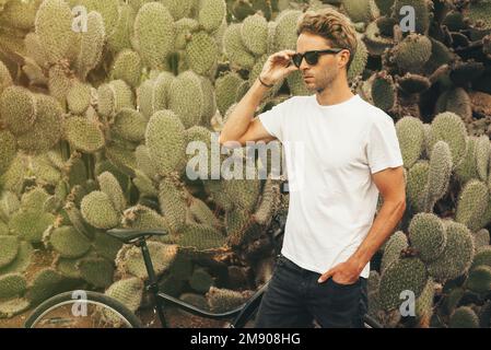 Blond hipster male model wearing sunglasses posing near a big cactus next to his bike. Stock Photo