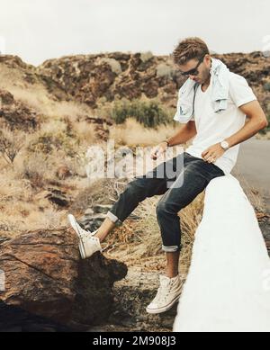 Young handsome guy sitting on parapet by the rocky hill wearing glasses, white blank t-shirt and black jeans. Stock Photo