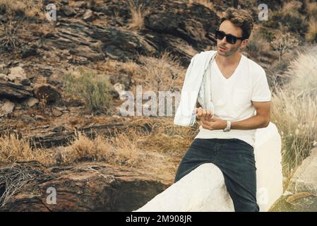 Young handsome guy sitting on parapet by the rocky hill wearing glasses, white blank t-shirt and black jeans. Stock Photo