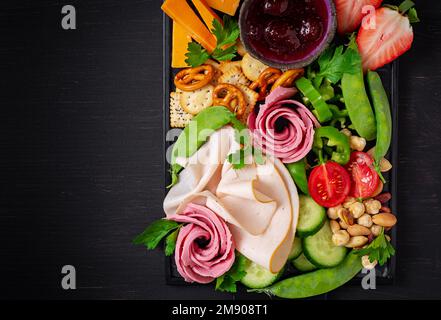 Antipasto platter cold sliced ham, salami, crackers, strawberries, vegetables and cheese platter on  board over dark background. Appetizers table with Stock Photo