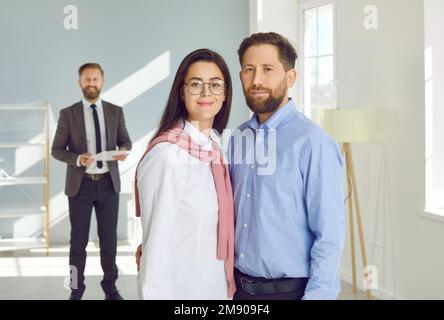 Portrait of a young couple standing in their new house, with an estate agent in the background Stock Photo