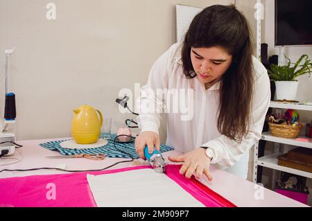 Caucasian young woman with long hair, working with the fabric cutter, tailoring a dress in her home sewing workshop Stock Photo