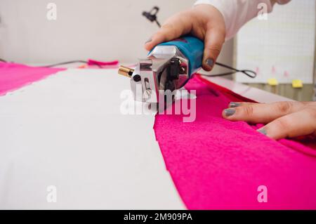 closeup female hands using of mechanical fabric cutter cutting fuchsia fabric, working from atelier at home, copy space, sewing and cutting concept Stock Photo