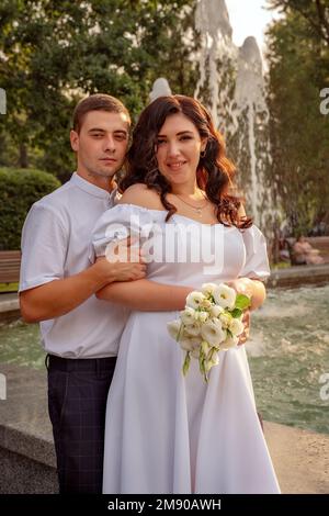 Woman in lush white dress poses against the backdrop of afountain in park, man in white shirt stands behind Stock Photo