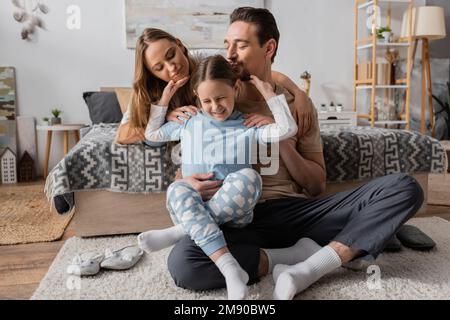 bearded man kissing head of cheerful kid sitting on carpet with dad near happy mother on bed,stock image Stock Photo