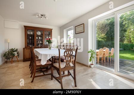 Sunlit dining room within modern house with traditional wooden furniture and sideboard, patio doors overlooking garden and white painted walls Stock Photo