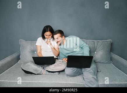 Young smiling couple relaxing on sofa with their laptops surfing the Internet, enjoying work from home. Stock Photo