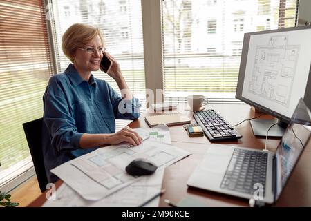 Mature woman engineer constructor talking phone with client while works on project from home office Stock Photo