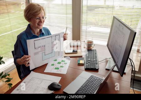 Mature smiling woman talking with client via online video call and showing project from home office Stock Photo