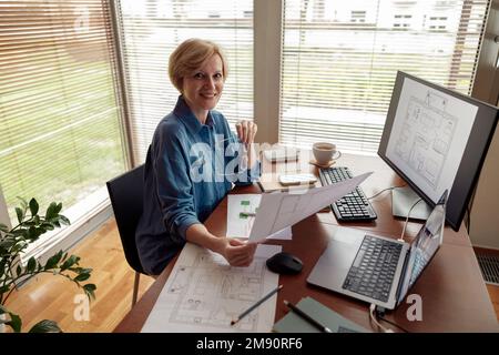 Smiling mature woman engineer constructor works on project from home office using computer Stock Photo