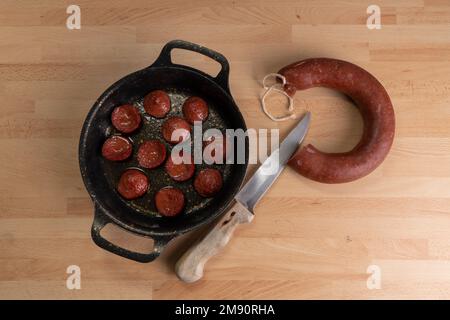 Top view of fried sausage slices in pan. Next to the frying pan are raw sausage and a knife on wooden background. Stock Photo