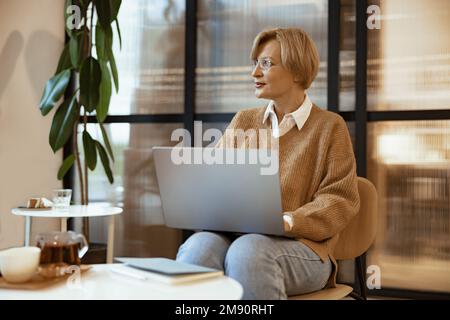 Smiling mature middle aged businesswoman using laptop working and web surfing in cozy coworking Stock Photo