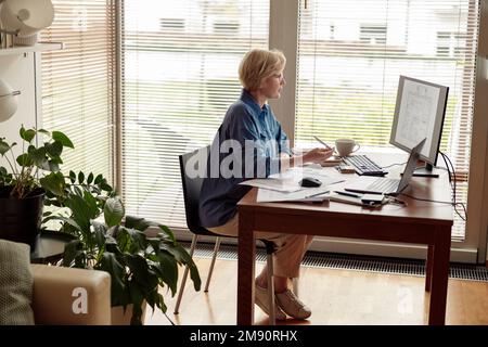 Focused mature woman engineer constructor works on project from home office using computer Stock Photo