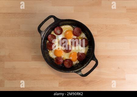 Top view of fried eggs with sausage slices in pan on wooden background. Stock Photo