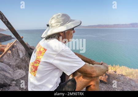 A healthy and fit 69-year-old male sits wearing a sun hat on the beach looking off into the Sea of Cortez, Baja, Mexico. Stock Photo