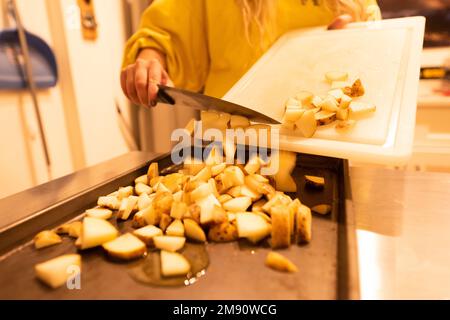 A closeup shot of a woman, putting chopped potatoes with a knife into an oily baking tray, in the kitchen, the concept of a meal preparation Stock Photo