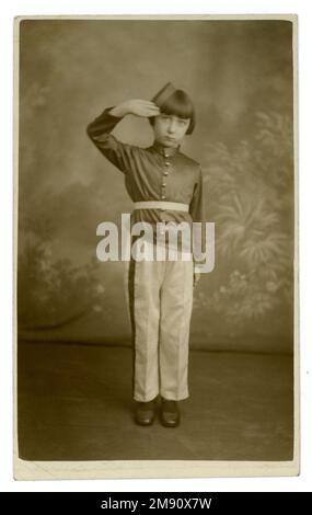 Original 1930's postcard of a cute young girl dressed in the uniform of a youth brass band, military marching band. 1930's childhood. 1930's girl. 1930's child. 1930's children. She wears silk trousers and tunic and bellhop hat and is saluting. Postcard is dated 6th Jan 1934 on reverse, studio of Williams Pioneers Studios Ltd. Tooting, Kingsland, Deptford, London. Stock Photo