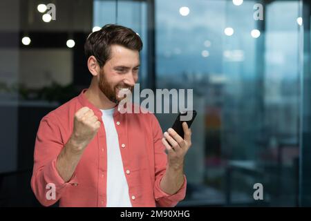 A successful businessman joyfully celebrates victory and good achievements, a man in a red shirt inside the office received an online notification, holding a smartphone in his hands Stock Photo
