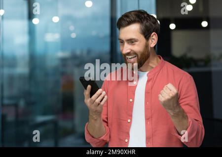 A successful businessman joyfully celebrates victory and good achievements, a man in a red shirt inside the office received an online notification, holding a smartphone in his hands Stock Photo