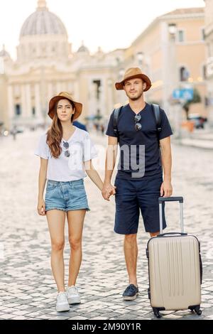 Happy young couple of travellers with hats and a suitcase hugging in the city and enjoying romance. Lovers smiling. A couple in love smiles and dreams Stock Photo