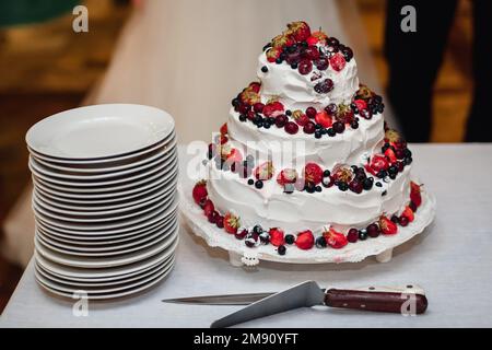 white cream wedding cake with fresh sweet strawberries, cherries, blueberries, black currants with three tiers on the table with plates Stock Photo