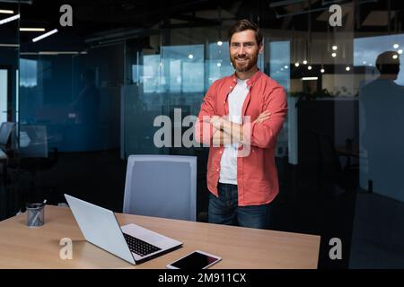 Portrait of a successful businessman inside the office, a man with crossed arms smiles and looks at the camera, an employee stands near a workplace with a computer in a red shirt and glasses. Stock Photo