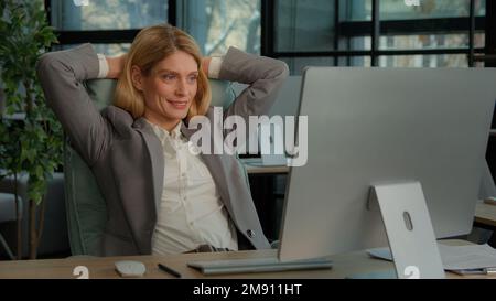 Middle-aged adult Caucasian woman 40s satisfied businesswoman finished computer work stretching sitting at workplace. Relaxed office worker female Stock Photo