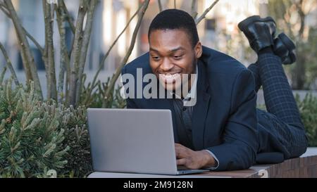 Smiling satisfied ethnic businessman corporate employee African American funny worker man lying down on bench work with laptop work communicate in Stock Photo