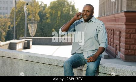 Adult mature African American man sitting outdoors holding cell phone angry middle-aged ethnic male businessman in city talking with smartphone has Stock Photo