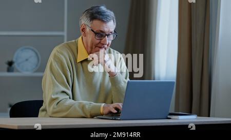 Old Caucasian man with computer work indoors. Elderly mature senior businessman worker entrepreneur grandfather at office at home at table with laptop Stock Photo