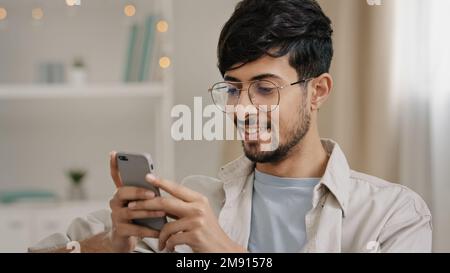 Young 30s arab bearded man millennial guy in glasses sits at home on couch looking at mobile phone making yes gesture getting offer winning good news Stock Photo