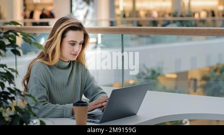 Caucasian business female freelancer student designer worker sitting at table in cafe office corporate space working with laptop typing message Stock Photo