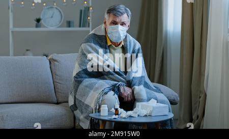 Upset sick grandpa in medical mask sitting on couch caucasian elderly man wrapped in blanket feel chills ill mature senior looking at camera symptoms Stock Photo