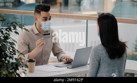 Man Hispanic bank worker in medical face mask consulting unrecognizable client woman about financial service debit easy payment banking account Stock Photo