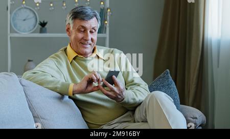 Caucasian elderly adult man sitting on sofa at home with gadget using online app on smartphone makes online order old grandfather resting reading news Stock Photo