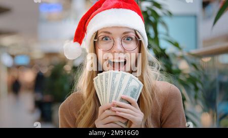 Close up rich Caucasian happy success woman lady in red Santa Christmas hat cap and eyeglass holding money dollars cash credit financial prize winning Stock Photo