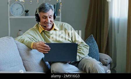 Mature Caucasian 60s talking man working distantly from home sitting on couch in headset talking to web camera laptop. Older 60s online teacher coach Stock Photo