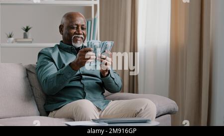 Happy old bald overweight African American senior mature elderly man happy with financial savings salary at home sitting couch. Winning online lottery Stock Photo