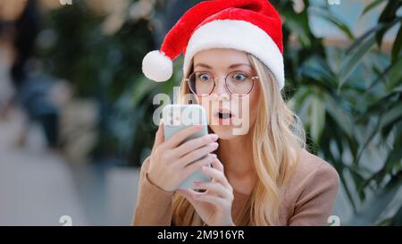 Surprised wonder astonish shocked amazed Caucasian woman girl lady in Santa Claus Christmas hat and eyeglass looking at phone mobile win victory offer Stock Photo