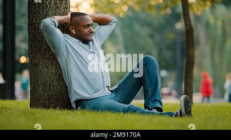 Happy calm relaxed elderly middle-aged mature senior adult African American ethnic man in wireless headphones sitting on grass in city park under tree Stock Photo