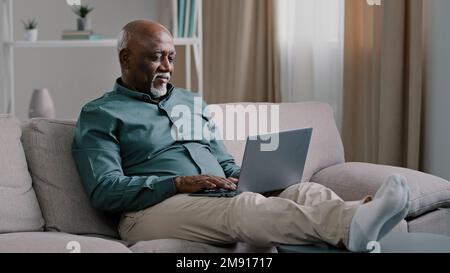 African bald overweight obese old middle-aged man senior businessman sitting on sofa at home working with laptop typing feels tired stress relaxing Stock Photo