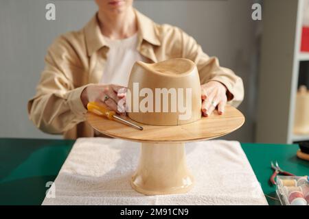 Close-up of wooden hat workpiece on table against young creative craftswoman working over new model for client of her studio Stock Photo