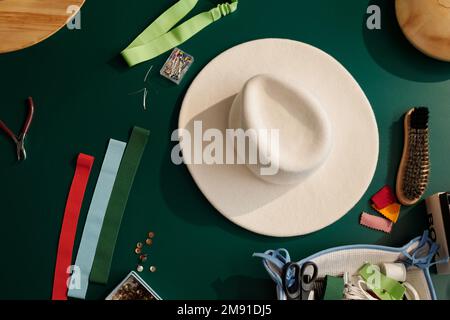Top view of white felt hat among variety of textile samples, pins in small box, brush for cleaning new items and other supplies on table Stock Photo
