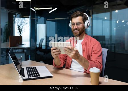 Successful businessman at workplace smiling man watching online video sitting at desk wearing headphones and glasses programmer holding tablet computer inside office. Stock Photo