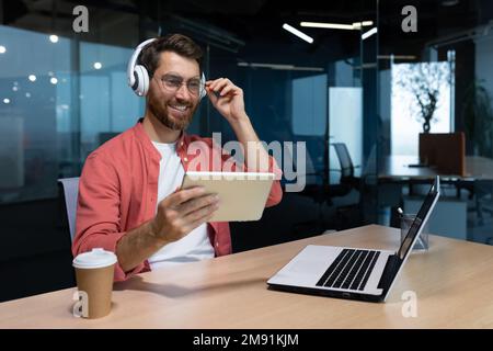 Successful businessman at workplace smiling man watching online video sitting at desk wearing headphones and glasses programmer holding tablet computer inside office. Stock Photo